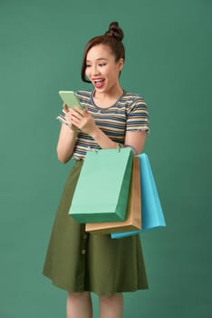 Cheerful young girl standing over green background, carrying shopping bags, using mobile phone