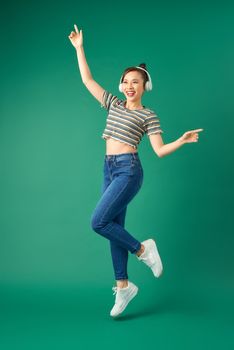 Full length portrait of young Asian woman in headphones listening to music dancing isolated green background