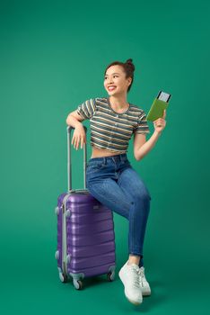Joyful young Asian girl sitting on suitcase and holding passport, flight ticket to travel over green background.