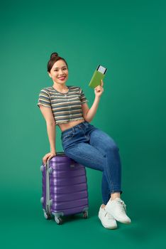 Joyful young Asian girl sitting on suitcase and holding passport, flight ticket to travel over green background.