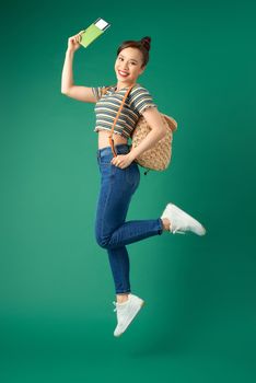 Beautiful young Asian girl traveler jumping in air while wearing bags and holding flight ticket, passport over green background.
