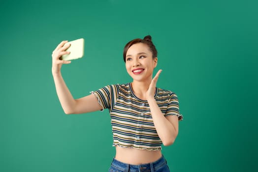 Portrait of young Asian female making selfie photo on smartphone with postitive expression over green background.