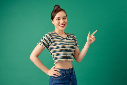 Happy young Asian woman in casual clothing pointing her finger over green background.