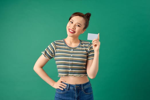 Portrait of cheerful young Asian woman showing credit card over green background.