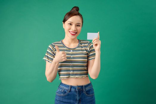 Smiling young Asian woman standing over green wall and holding debit card in hands.