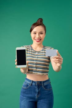 Happy young Asian woman showing blank mobile phone and credit card isolated over green background.