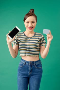 Happy young Asian woman showing blank mobile phone and credit card isolated over green background.