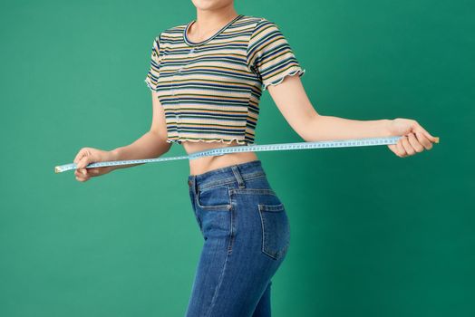 Close up young woman measuring her waist with tape measure over green background.