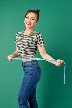 Beautiful young Asian woman measuring her waist with a measure tape over green background.