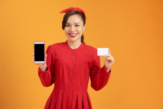 happy young Asian woman shows blank credit card and smart phone over orange background.