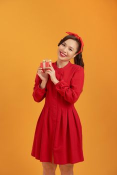 Cheerful young Asian woman holding gift box when standing over orange background.