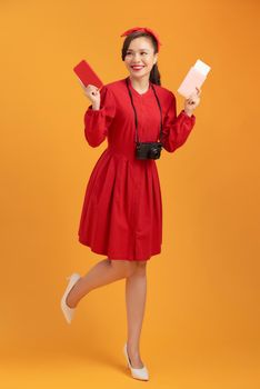 Happy young Asian woman tourist holding phone, passpoer, camera and standing over orange background.