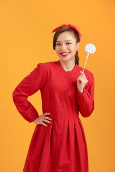 Smiling young Asian woman in red dress holding lollipop while standing over orange backgound.