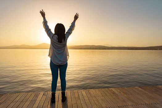 Silhouette of brunette girl view from back in transparent shirt standing on a pier with open arms up to the sky looking at horizon with setting sun with reflection on sea ocean water. Pensive concept