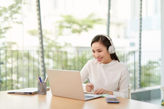 Smiling young woman sitting at the kitchen, using laptop computer for study, listening to music with headphones