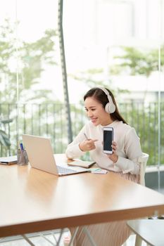 Video conference, video meeting. A young woman works from home, she uses a headset for online communication. She points finger to the phone screen. Work remotely from home