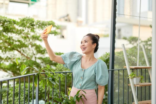 Cheerful young Asian girl having a video call with her phone when standing o balcony.