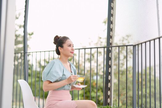 A smiling girl is drinking tea while sitting at a table on the open balcony. Rest and happiness concept
