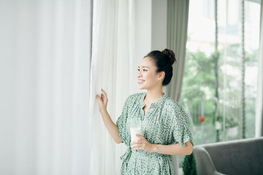 Morning portrait of young smiling woman drinking milk from the glass, woman standing at home near the window, healthy food eating.
