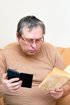 Senior citizen sitting on the couch understands the gadget, mastering the smartphone. Pensioner holds a smartphone and a book