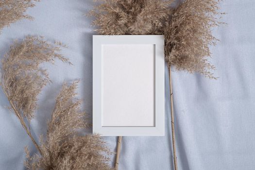 Picture frame with blank paper card mockup near to pampas dry grass on blue neutral colored textile
