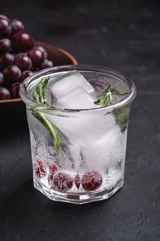 Fresh ice cold carbonated water in glass with rosemary leaf near to wooden bowl with grape berries, dark stone background, angle view