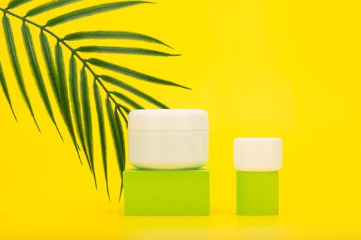 Face cream or mask and lip balm, scrub or under eye cream on green podiums against yellow background with palm leaf and copy space. Concept of beauty products with sunscreen and summer skin care 