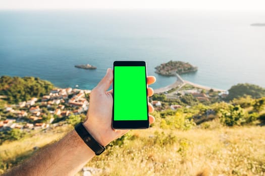 Man's hand holds a smartphone with a blank screen vertically on the background of Sveti Stefan island Montenegro. High quality photo