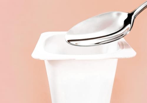 Yogurt cup and silver spoon on beige background, white plastic container with yoghurt cream, fresh dairy product for healthy diet and nutrition balance.