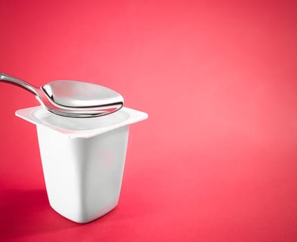 Yogurt cup and silver spoon on red background, white plastic container with yoghurt cream, fresh dairy product for healthy diet and nutrition balance.