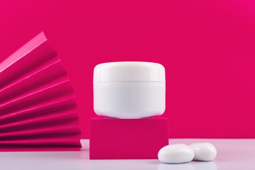 White cosmetic jar on pink podium with two white stones next to it against pink background decorated with waver. Concept of skin care product, face cream, gel, mask or scrub