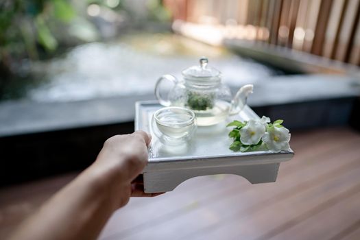Hand holding the tray served transparent teapot with herbal tea.