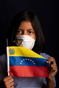 Portrait of little Venezuelan girl wearing medical mask and holding hopefully the flag of Venezuela