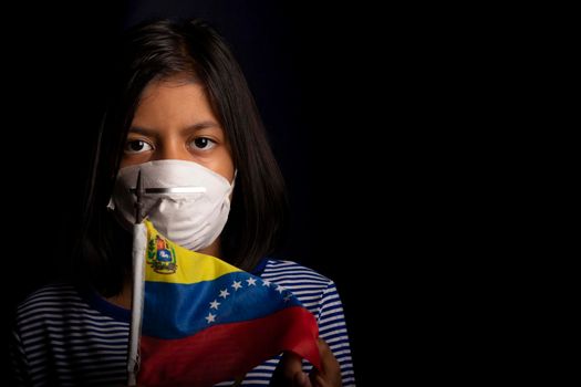 Portrait of little Venezuelan girl wearing medical mask and holding hopefully the flag of Venezuela