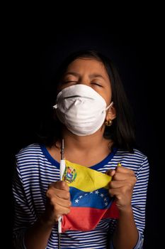 Portrait of little Venezuelan girl wearing medical mask and holding hopefully the flag of Venezuela