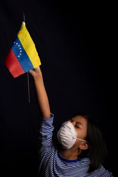 Portrait of little Venezuelan girl wearing medical mask and holding hopefully the flag of Venezuela