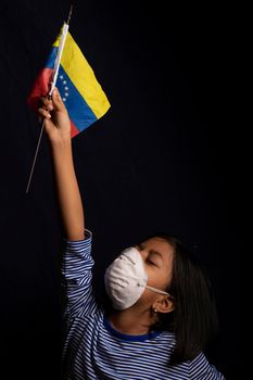 Portrait of little Venezuelan girl wearing medical mask and holding hopefully the flag of Venezuela