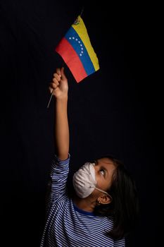 Portrait of little Venezuelan girl wearing medical mask and holding hopefully the flag of Venezuela