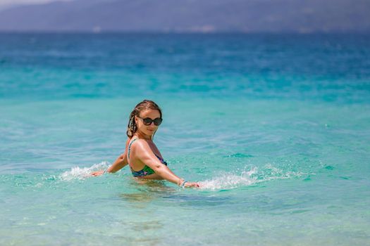 Young beautiful girl splashes in the blue water of the ocean. Rejoices and laughs on vacation