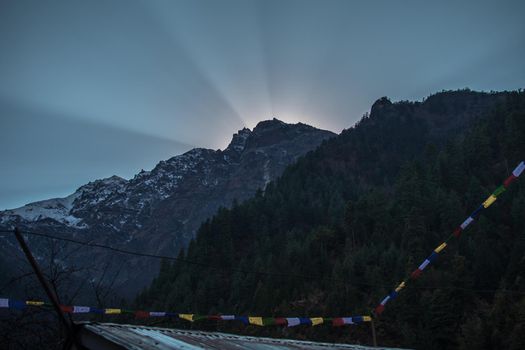 Bright sun rays peaking over the nepalese mountain tops over buddhist prayer flags