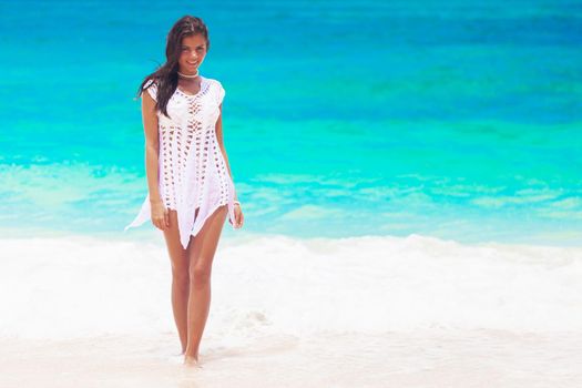 Happy young woman wearing white dress is walking on the beach of tropical sea and smiling