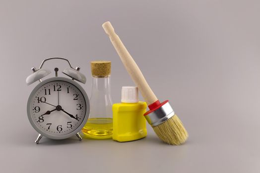 New color, decorating or renovations concept with a still life of an alarm clock with clean paint brush, unlabeled yellow pigment container and laboratory flask over a grey background