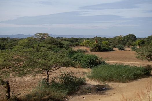 BAGAN, NYAUNG-U, MYANMAR - 3 JANUARY 2020: Several old and historical temple pagodas and green vegetation in the distance from an open dry grass and hay field