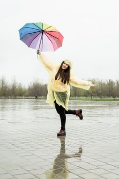 Beautiful brunnette woman in yellow raincoat holding rainbow umbrella dancing out in the rain