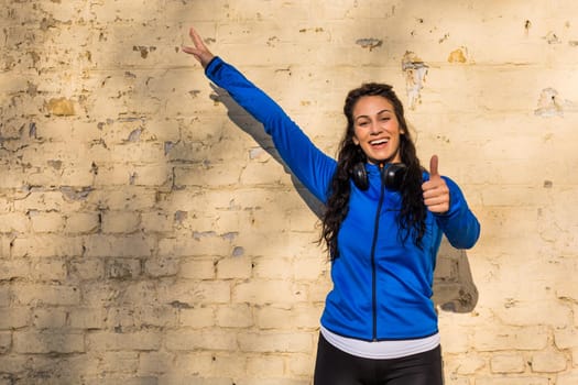 Portrait of happy sporty woman showing thumb up while standing in front of wall .
