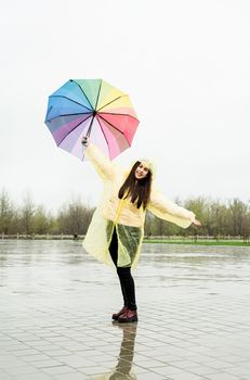 Beautiful brunette woman in yellow raincoat holding rainbow umbrella out in the rain