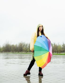 Beautiful brunette woman in yellow raincoat holding rainbow umbrella out in the rain, making funny face showing tongue out