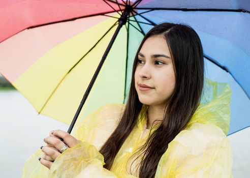 Beautiful smiling brunette woman in yellow raincoat holding rainbow umbrella out in the rain