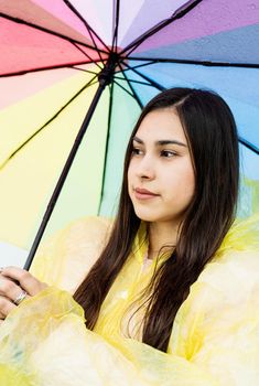 Beautiful smiling brunette woman in yellow raincoat holding rainbow umbrella out in the rain