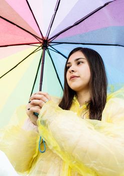 Beautiful smiling brunette woman in yellow raincoat holding rainbow umbrella out in the rain